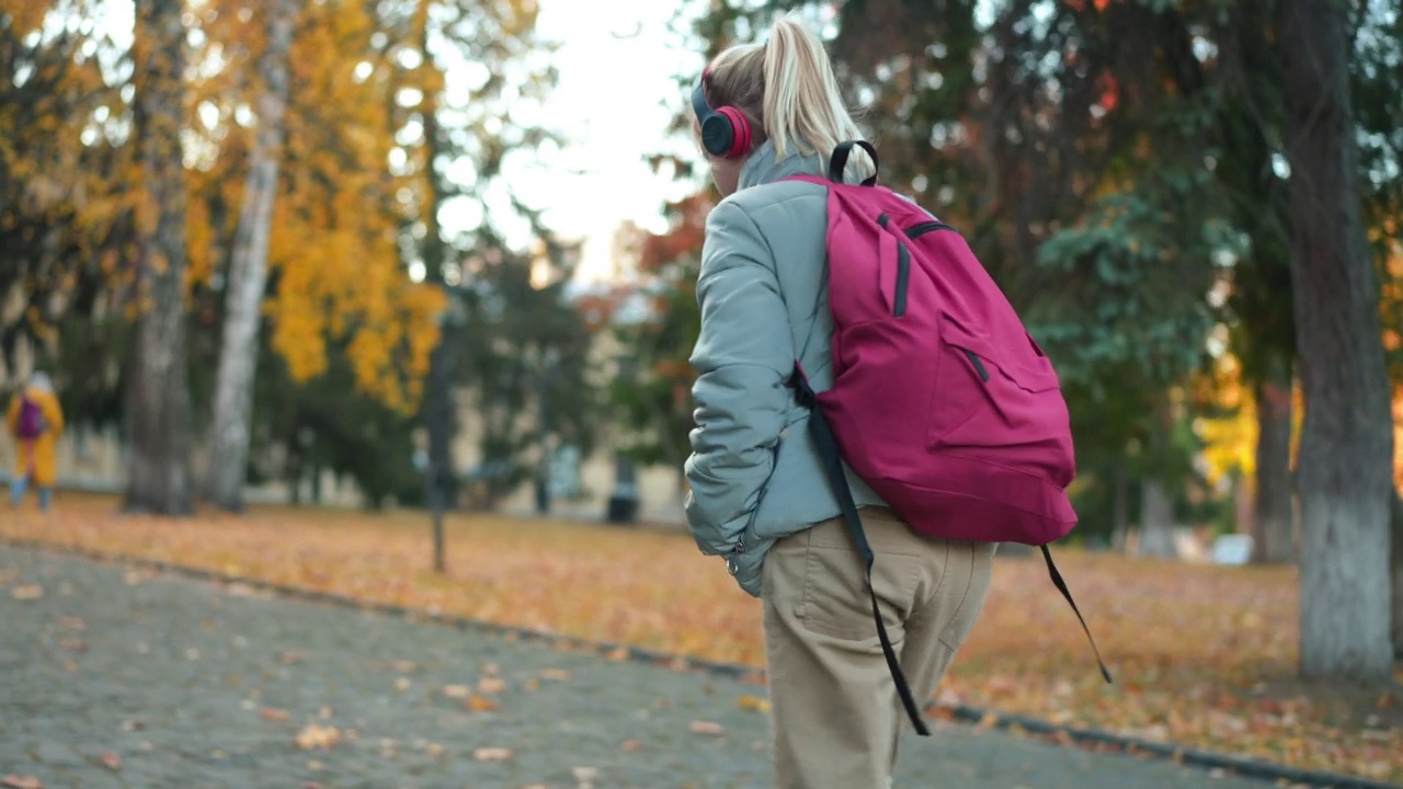 直播镜头跟随自信的少女戴着耳机走在校园里听音乐。跟踪拍摄的白人女孩漫步到大学在秋天的一天。爱好和教育。视频素材