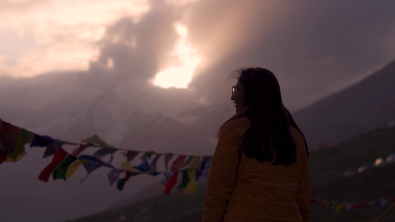 美丽的印度女孩旅行者的剪影站在雪山和经幡飘扬的风在日落。引人注目的日落天空在山上。女性享受假期视频素材