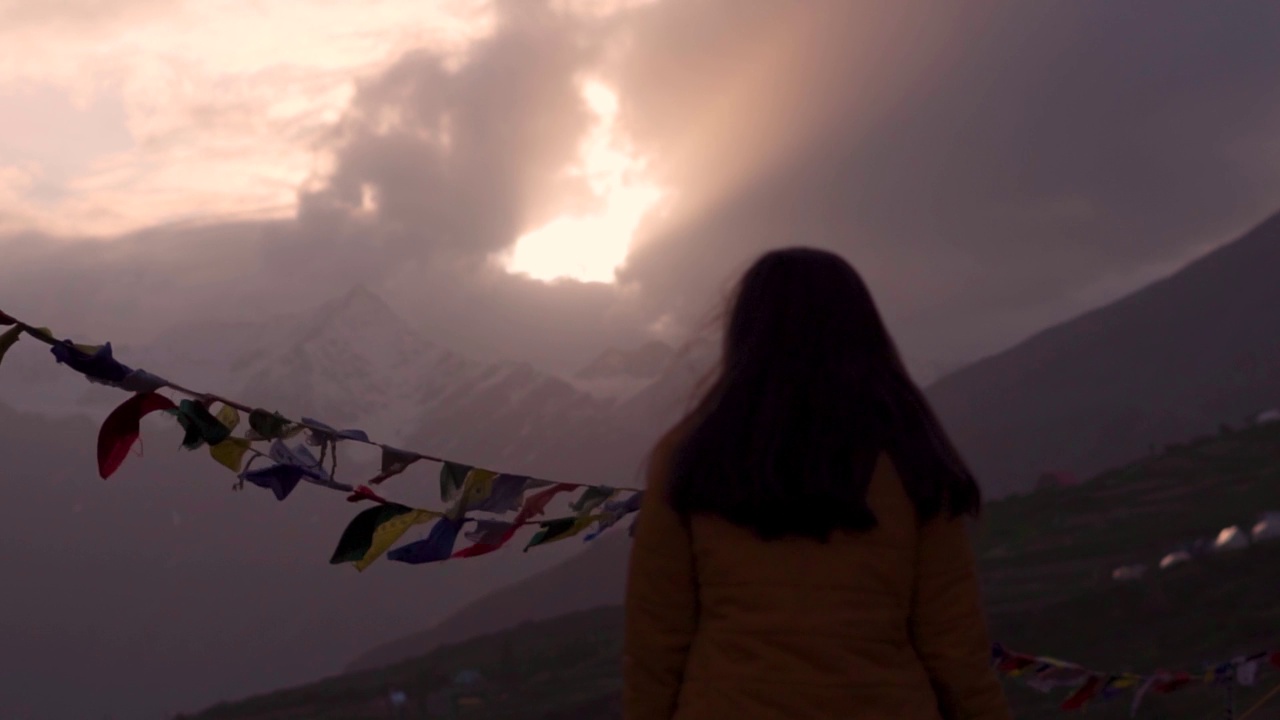 年轻的印度女旅行者在印度拉豪尔和斯皮提的喜马拉雅山脉欣赏美丽的日落。多云的日落天空和山与佛教经幡飘扬在风中。架的焦点视频素材