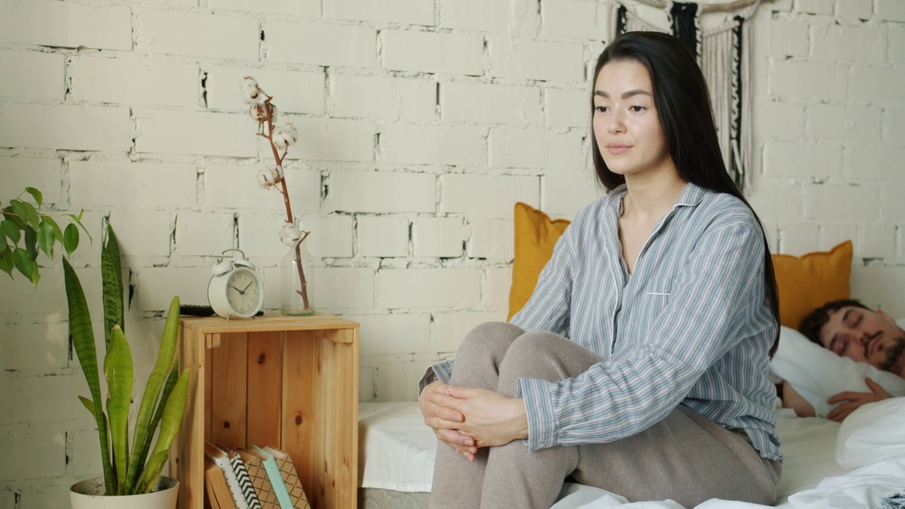 在家里的卧室里，沉思的亚洲女人坐在床上看着睡着的丈夫叹息视频素材