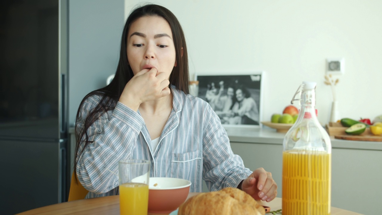 快乐的亚洲女性在家里的厨房里吃着羊角面包，喝着果汁，用着智能手机视频素材