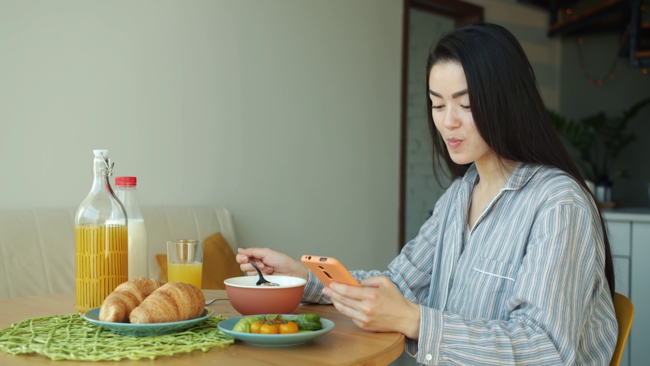 亚洲女性在家厨房吃早餐和使用智能手机的肖像视频素材