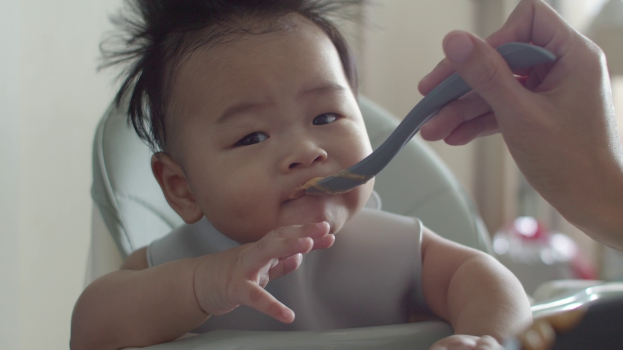 坐在高椅子上的亚洲小男孩正在被妈妈喂食。视频素材