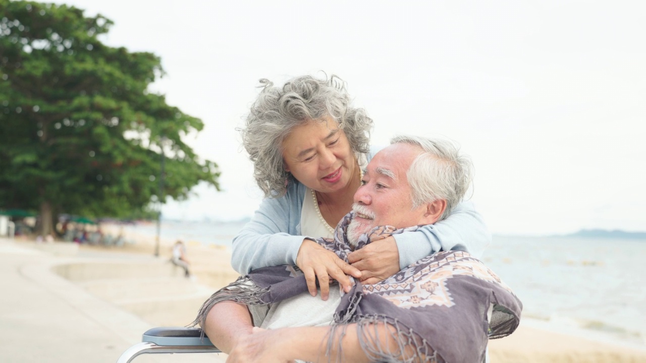 亚洲夫妇退休的老男人和女人坐在轮椅上放松在海滩上的周末假期与家人。爱与健康的理念。视频素材