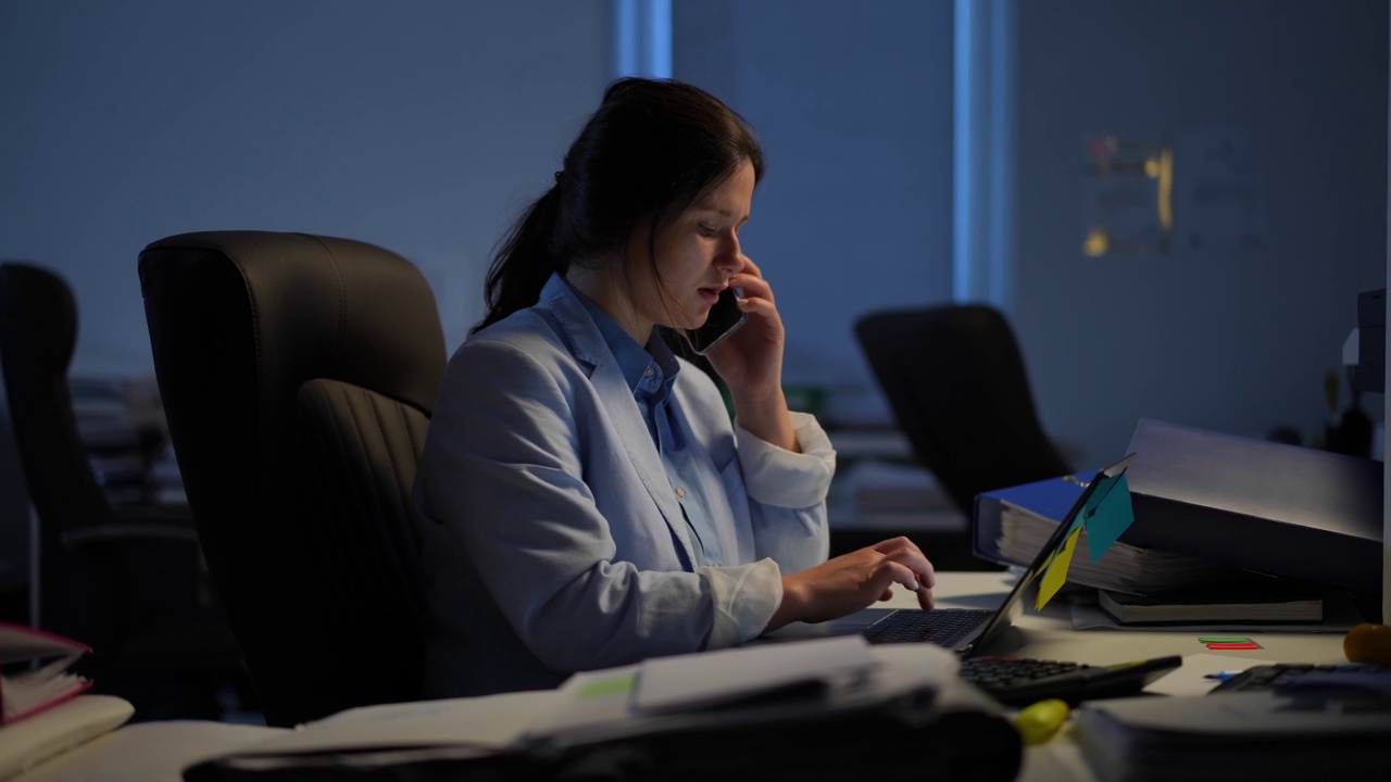 聪明漂亮的白种人女人上网，手提电脑接电话。集中自信的女性员工的侧视图肖像在办公室室内工作在晚上。视频素材