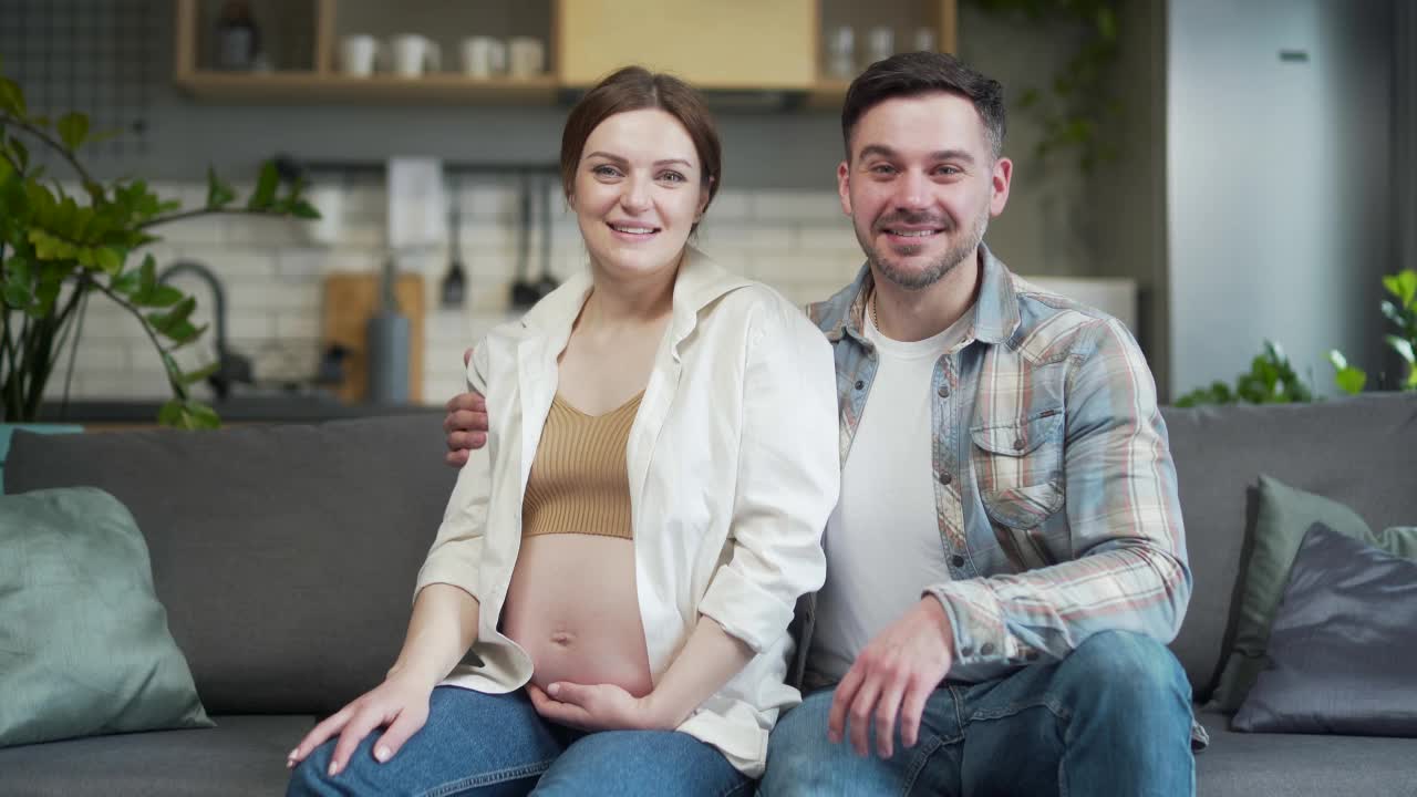 年轻的幸福夫妇通过视频电话在网上聊天。怀孕的妻子和丈夫一起对着镜头秀恩爱。坐在家里的沙发上。视频素材