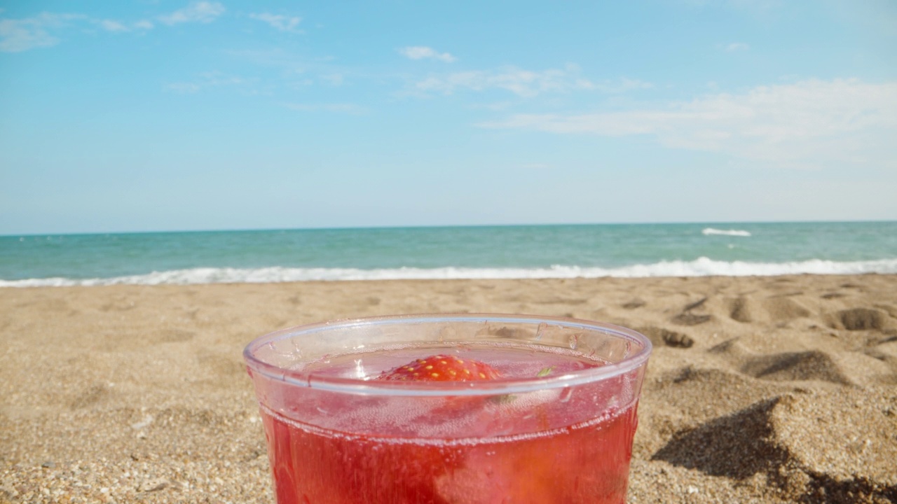 红色鸡尾酒在海滩上，以大海为背景，草莓在玻璃杯中旋转。特写镜头。视频素材