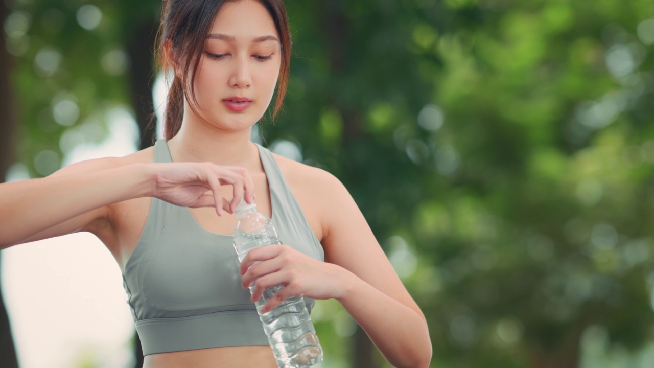 在公园慢跑时，穿着运动服喝瓶装水的快乐亚洲年轻女子。健康的女运动员喜欢运动训练、锻炼、晨跑视频素材