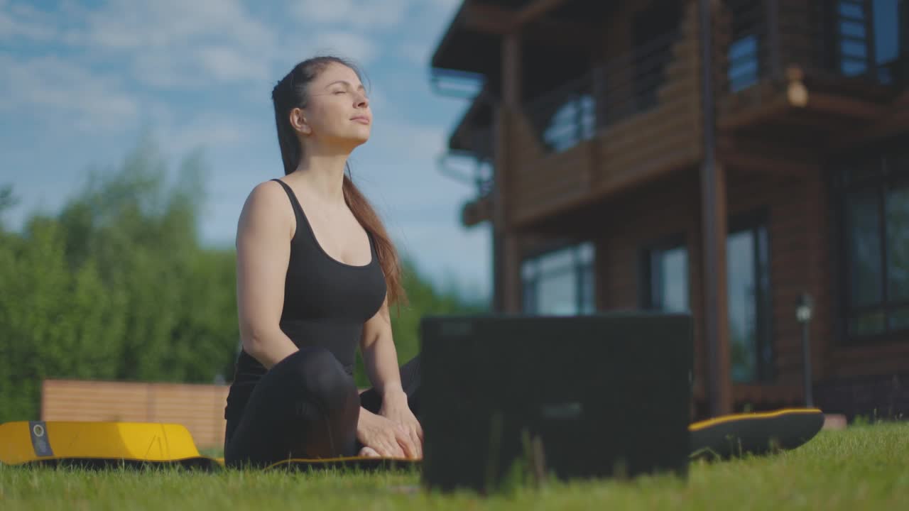 年轻专注的女孩坐在健身垫在笔记本电脑前莲花位置。这个女孩做瑜伽。慢动作视频素材