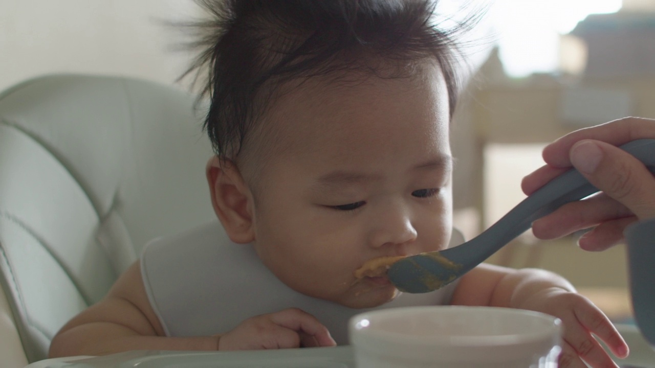 坐在高椅子上的亚洲小男孩正在被妈妈喂食。视频素材