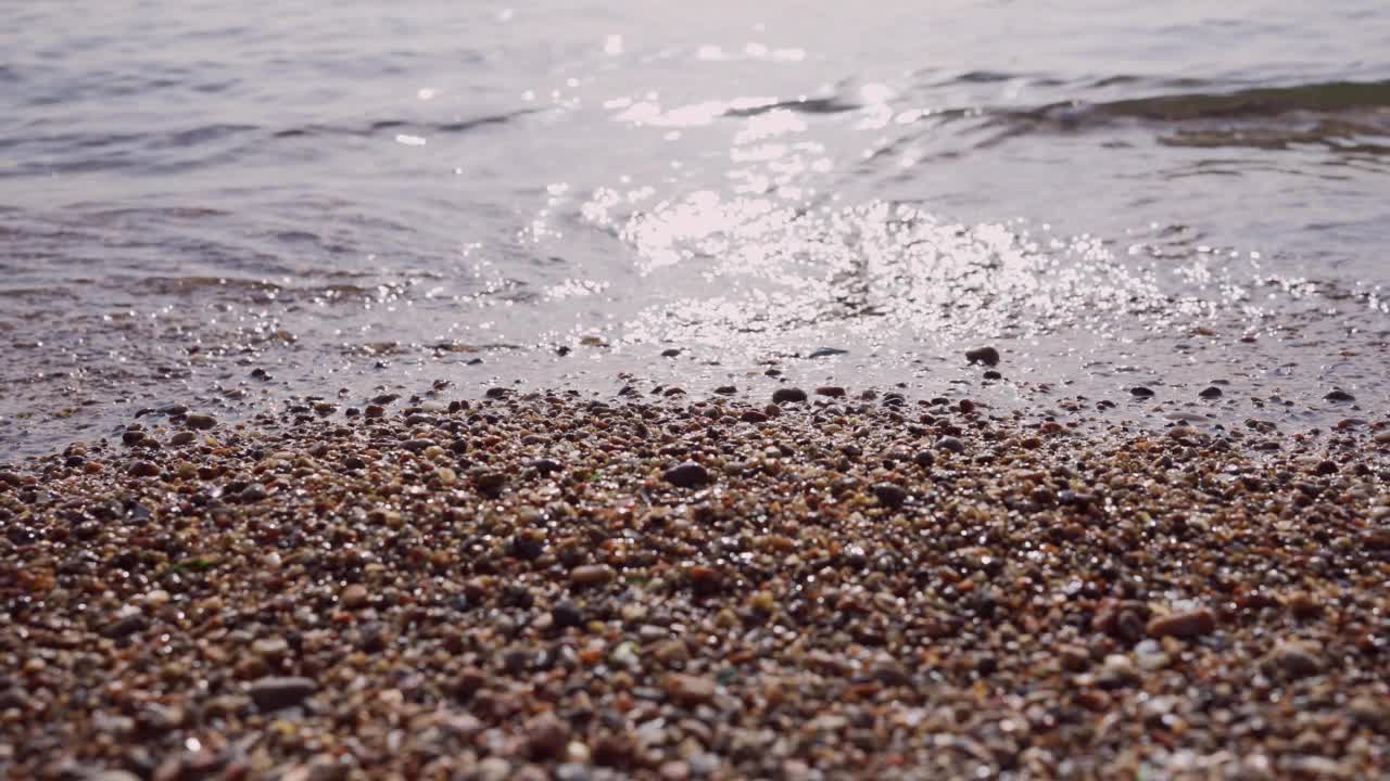 卵石滩的特写，透明的波浪，阳光刺眼的水面。靠近多石的海岸。海浪带平静大海的自然背景。相机向右侧移动视频素材