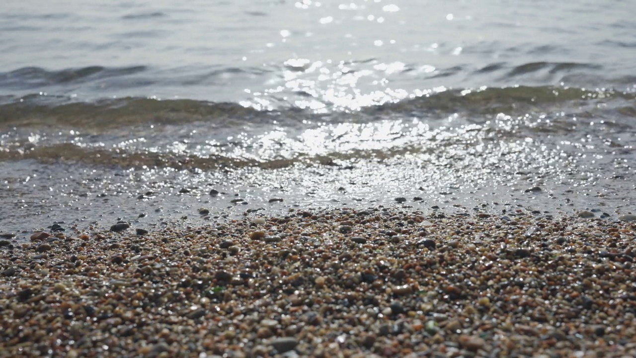 特写的卵石海滩与透明的波浪与阳光在水中眩光。近距离的石头海岸。冲浪区的自然背景是平静的大海。镜头移到左边视频素材