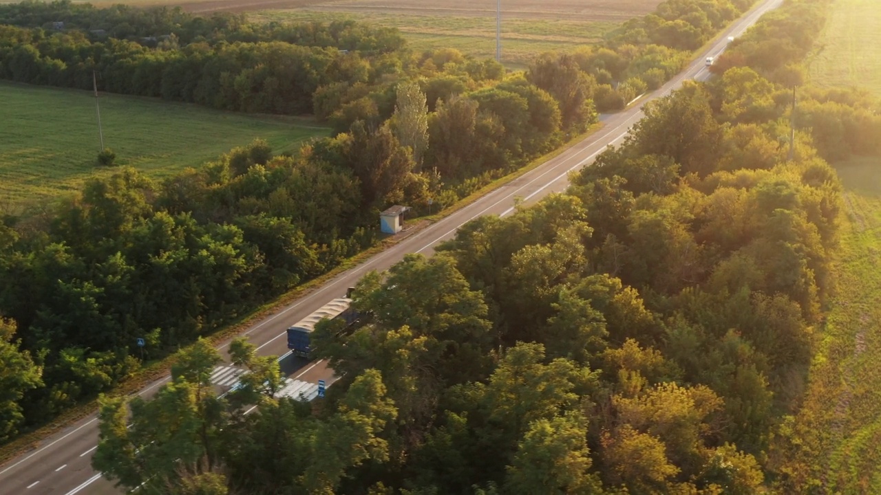 航拍的一辆谷物卡车在夏日夕阳下行驶在美丽的乡村道路上。无人机拍摄的卡车行驶在乡间的高速公路上，沿途有农田和树木。视频素材