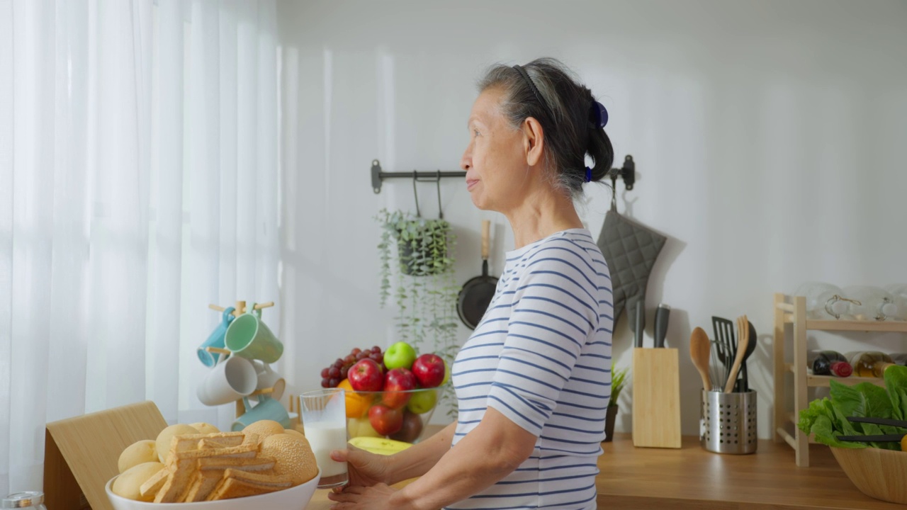 亚洲高级成熟女性在家里的厨房喝一杯牛奶。有魅力的老祖母抱着和喝着一杯牛奶，早上醒来在家里的保健。视频素材