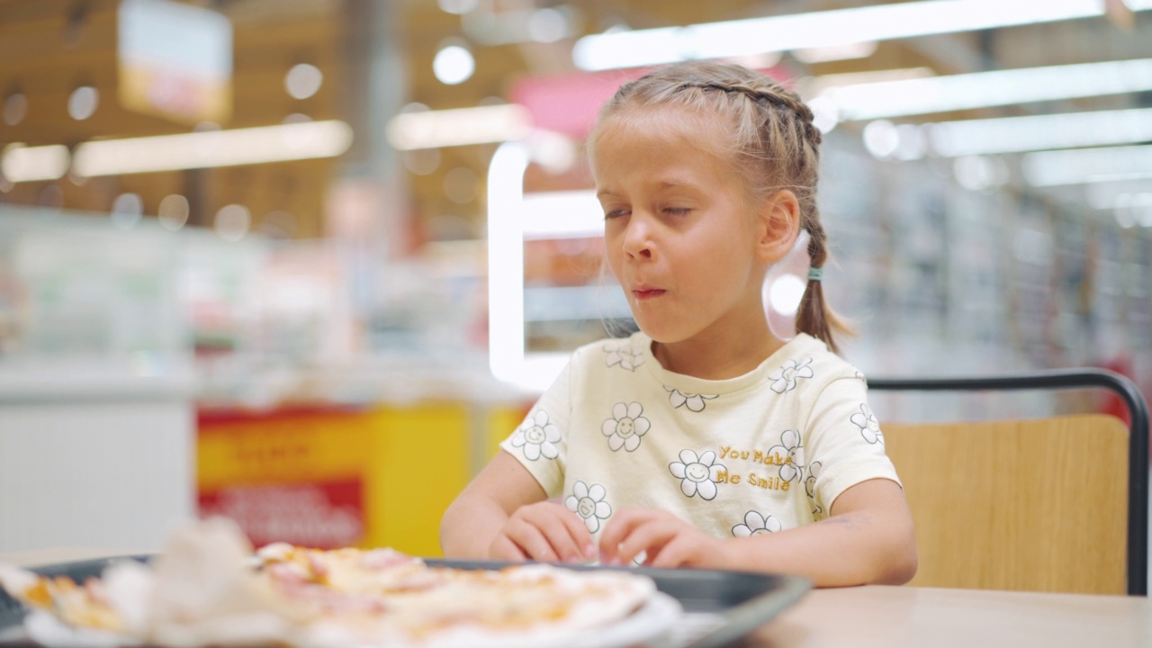 孩子坐在超市食品广场的桌子上吃披萨。小女孩在室内享受快餐营养。饥饿的孩子在吃披萨。肖像拍摄。视频素材