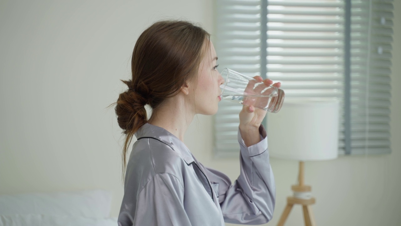 穿着睡衣的年轻白人妇女在厨房里喝一杯水。迷人的美丽口渴的女孩抱着清洁的天然矿物在杯子醒来后，在早上的健康护理在家里啜饮。视频素材