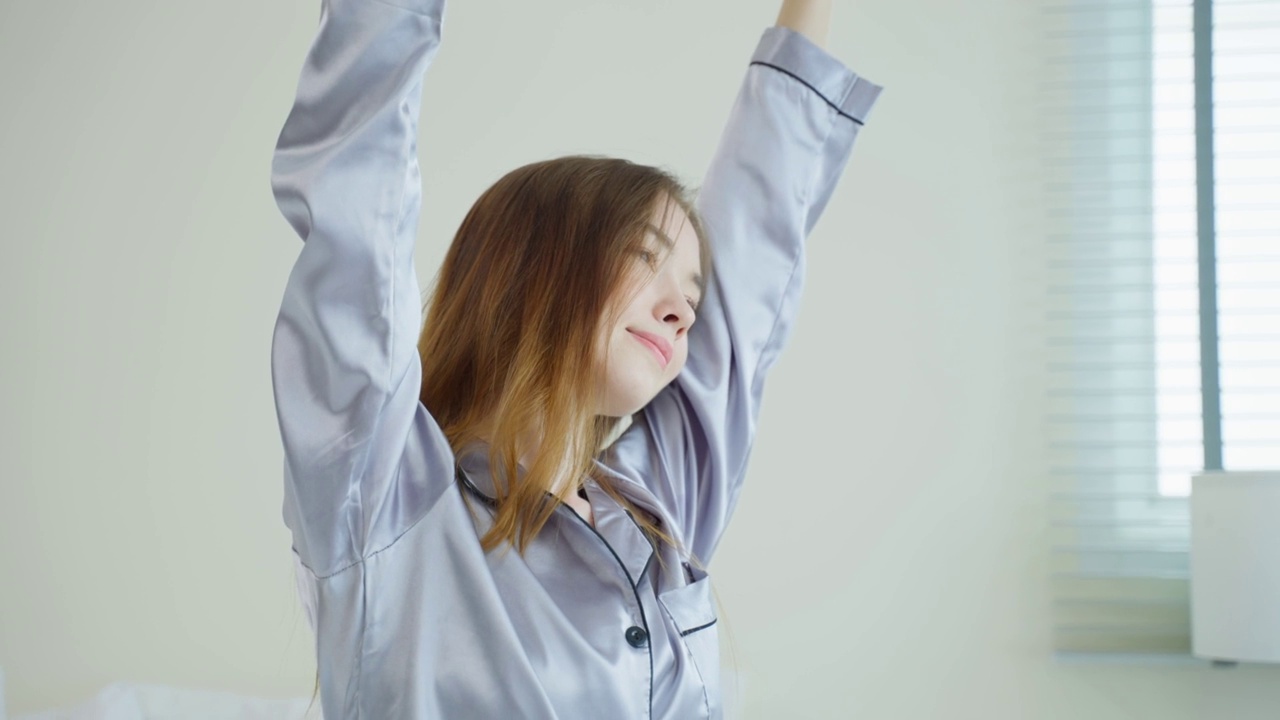 穿着睡衣的漂亮白人女孩早上醒来伸懒腰。有魅力的年轻女子微笑着，感到高兴和放松，然后伸展身体从床上睡觉在家里的卧室。视频素材