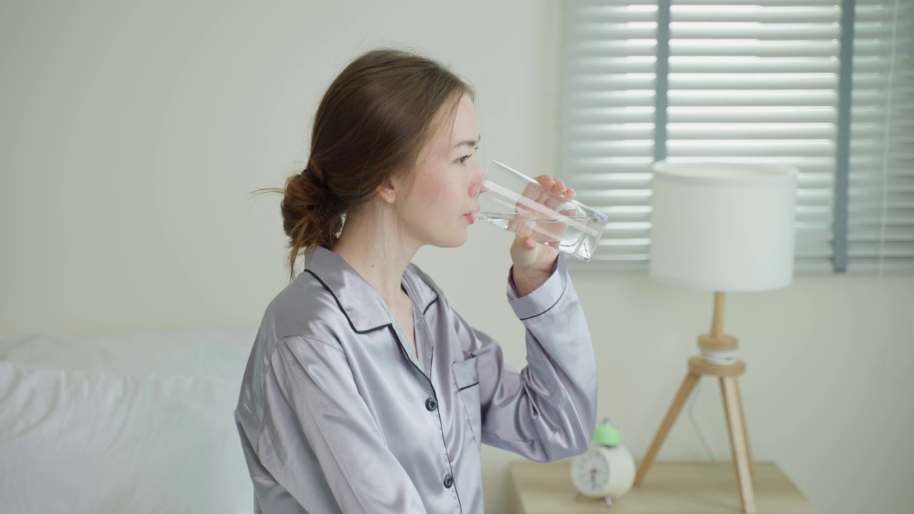 穿着睡衣的年轻白人妇女在厨房里喝一杯水。迷人的美丽口渴的女孩抱着清洁的天然矿物在杯子醒来后，在早上的健康护理在家里啜饮。视频素材