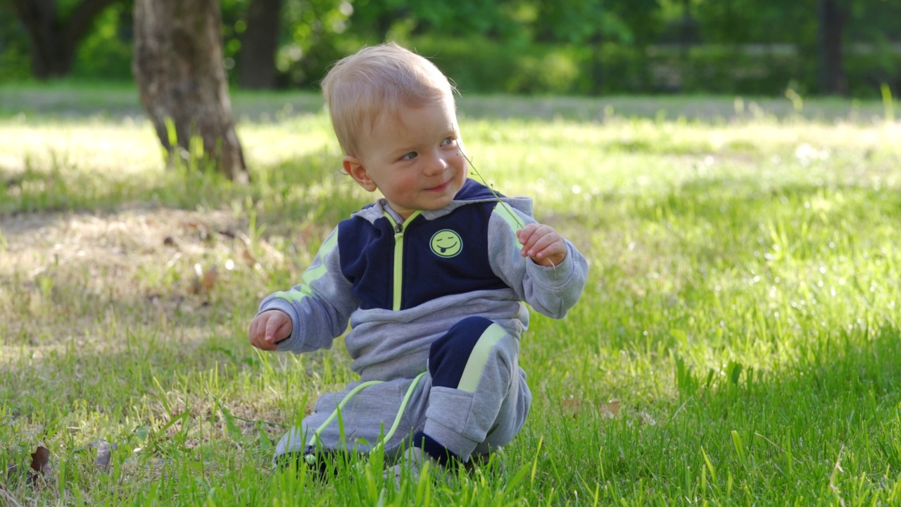 孩子坐在春天公园的绿草地上，1.5岁的幼童在绿草地上玩耍。儿童健康的夏季活动。视频素材