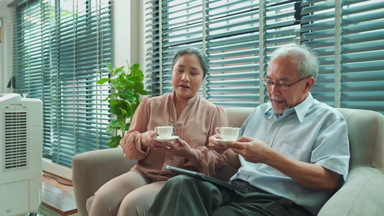 退休的亚洲老人坐在沙发上用平板电脑，老妇人给他端来一杯茶视频素材