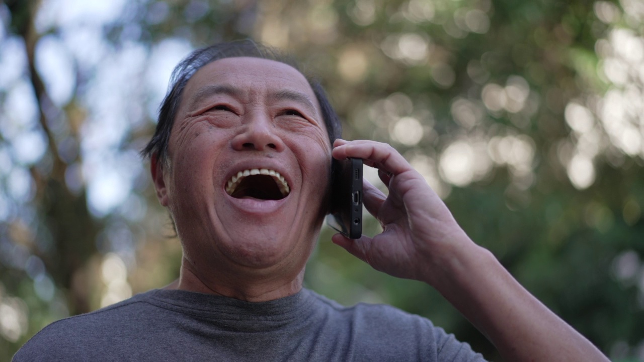 快乐的资深亚洲人讲电话，而走在城市的街道。一个中年老人的肖像在与手机设备交谈视频素材