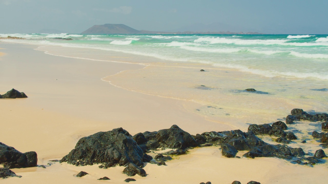 Fuerteventura,加那利群岛。西班牙。旅游娱乐。海浪冲过黑色的石头，白色的沙子冒着泡沫。自然、环保、自然奇观的理念。静态的镜头。视频素材