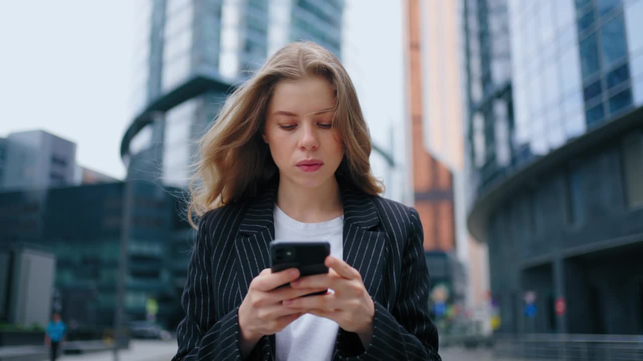 美丽的年轻女子使用智能手机穿过城市街道。微笑的女性肖像使用手机，发布社交媒体，网上购物，短信视频素材