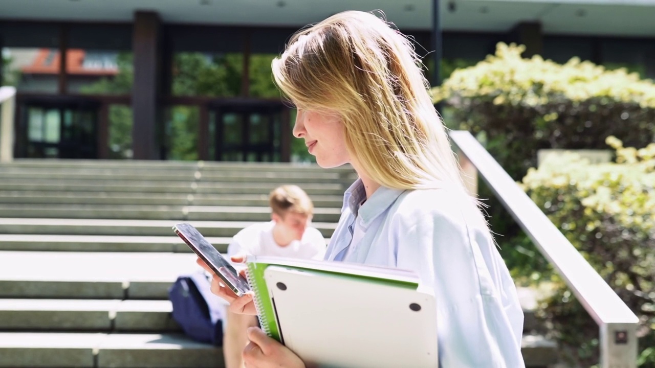 漂亮的女孩学生拿着手机用智能手机走在户外。视频素材