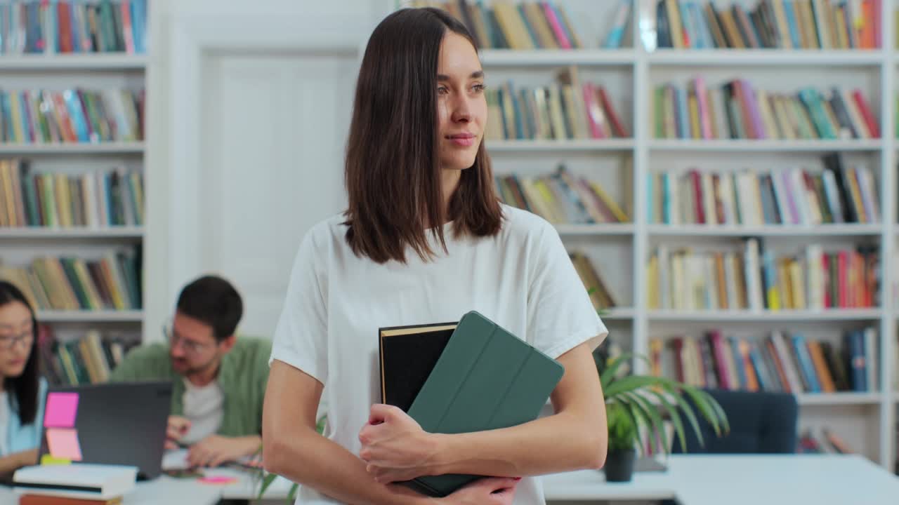 美丽的年轻女孩的肖像拿着书和数字平板电脑在图书馆的书架背景看着相机。大学教育的概念。视频素材