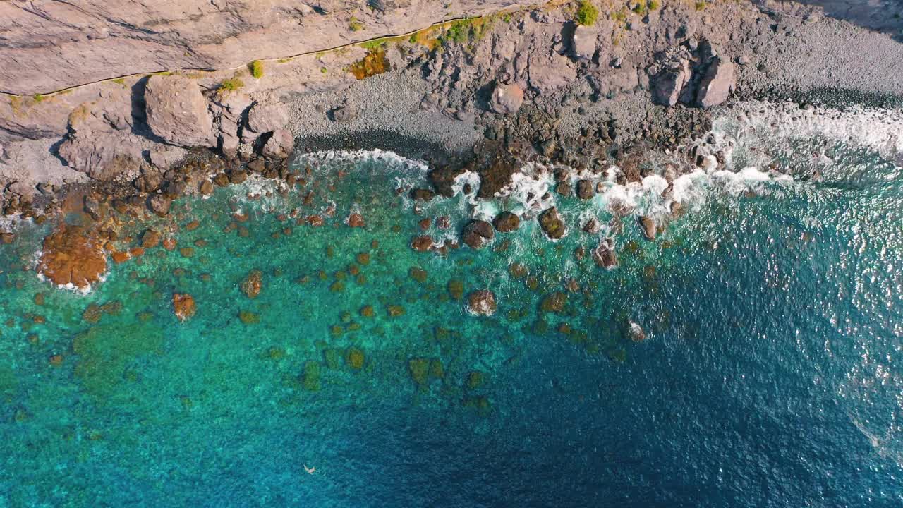 从上空俯视，海浪拍打着空旷的热带岩石海滩，上面有火山形成的岩石。阳光明媚的一天。狂野的海滩会有深蓝色湛蓝清澈的海水。视频素材