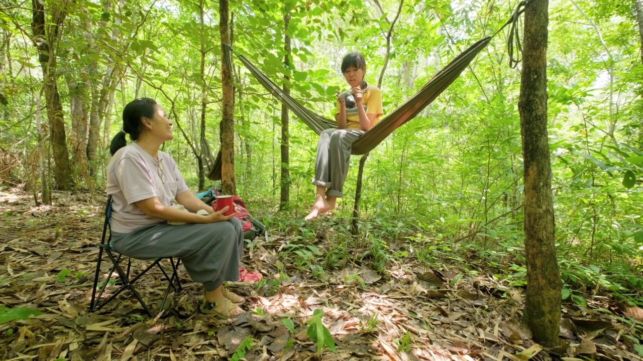 亚洲母女背着双肩包徒步旅行，一起在森林小径露营，在吊床上享受放松视频素材