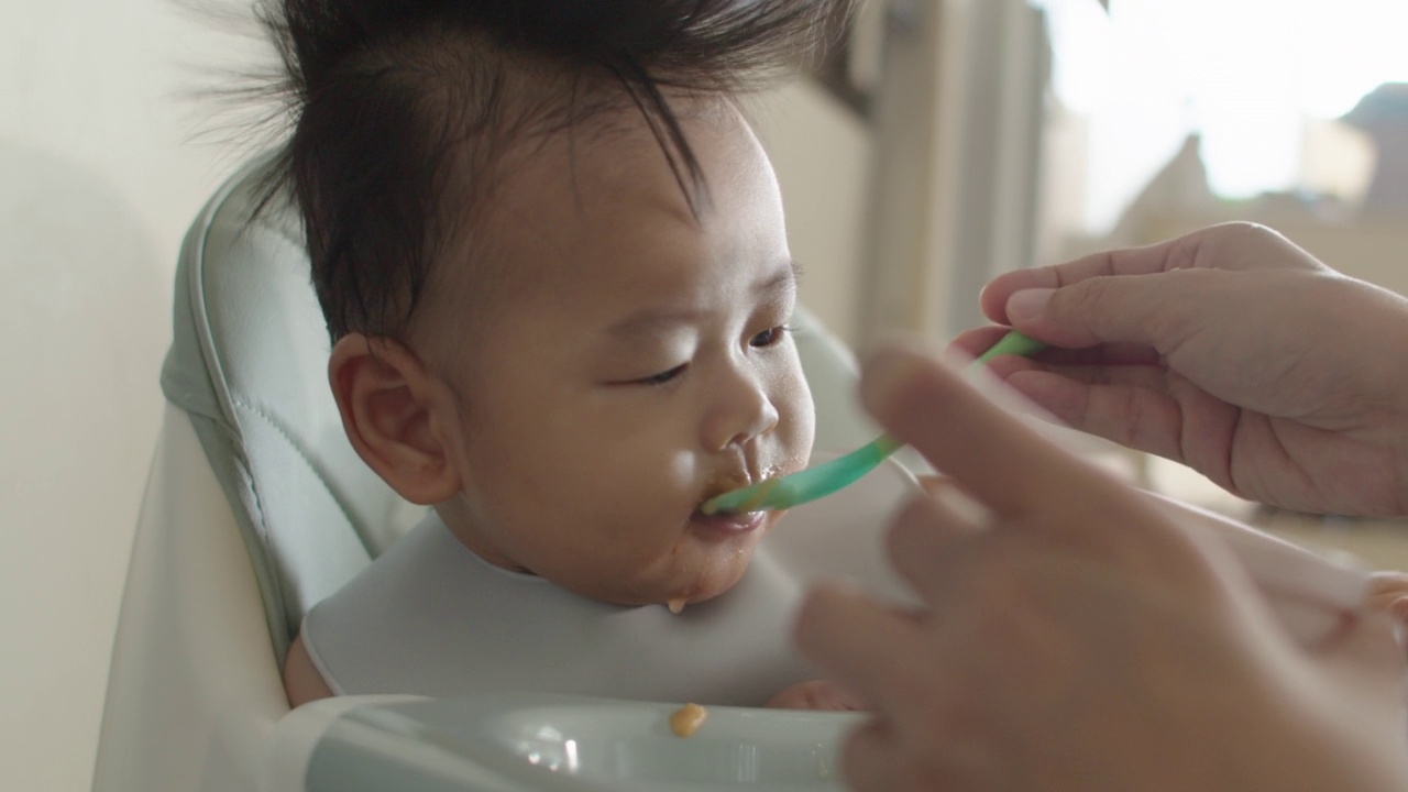 坐在高脚椅上的亚洲小男孩正在被妈妈喂食物。视频素材