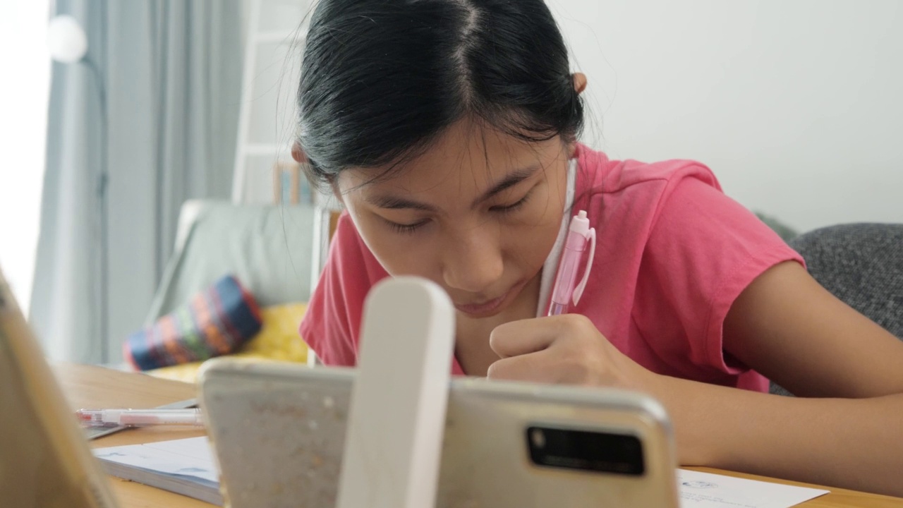 亚洲女孩一边做作业，一边在家用智能手机或平板电脑学习在线课程，保持社交距离的生活方式理念。视频素材