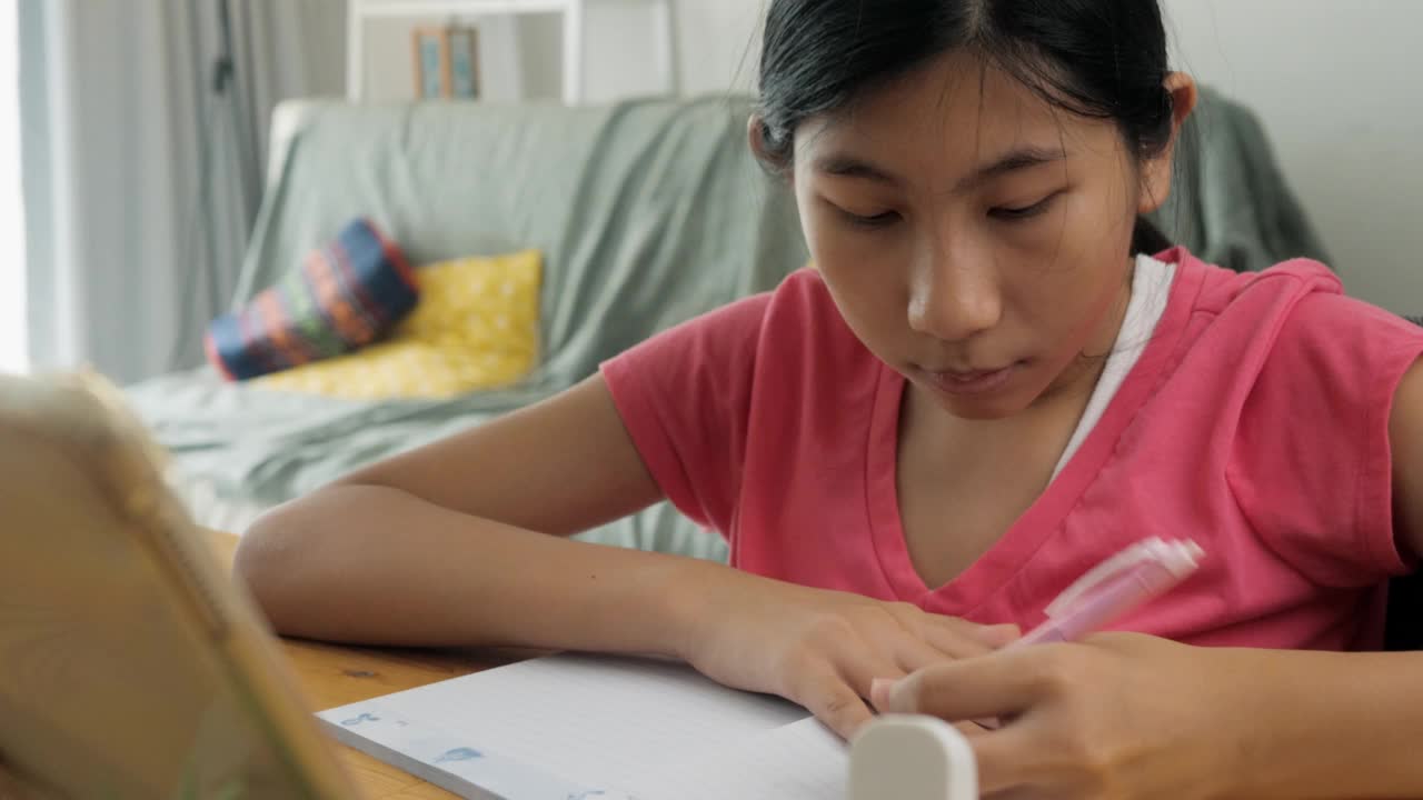 亚洲女孩一边做作业，一边在家用智能手机或平板电脑学习在线课程，保持社交距离的生活方式理念。视频素材