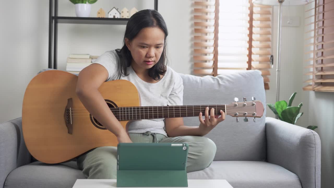 亚洲妇女坐在沙发上练习弹木吉他从网上教程在家里学习。以技能提升为理念的周末活动视频素材