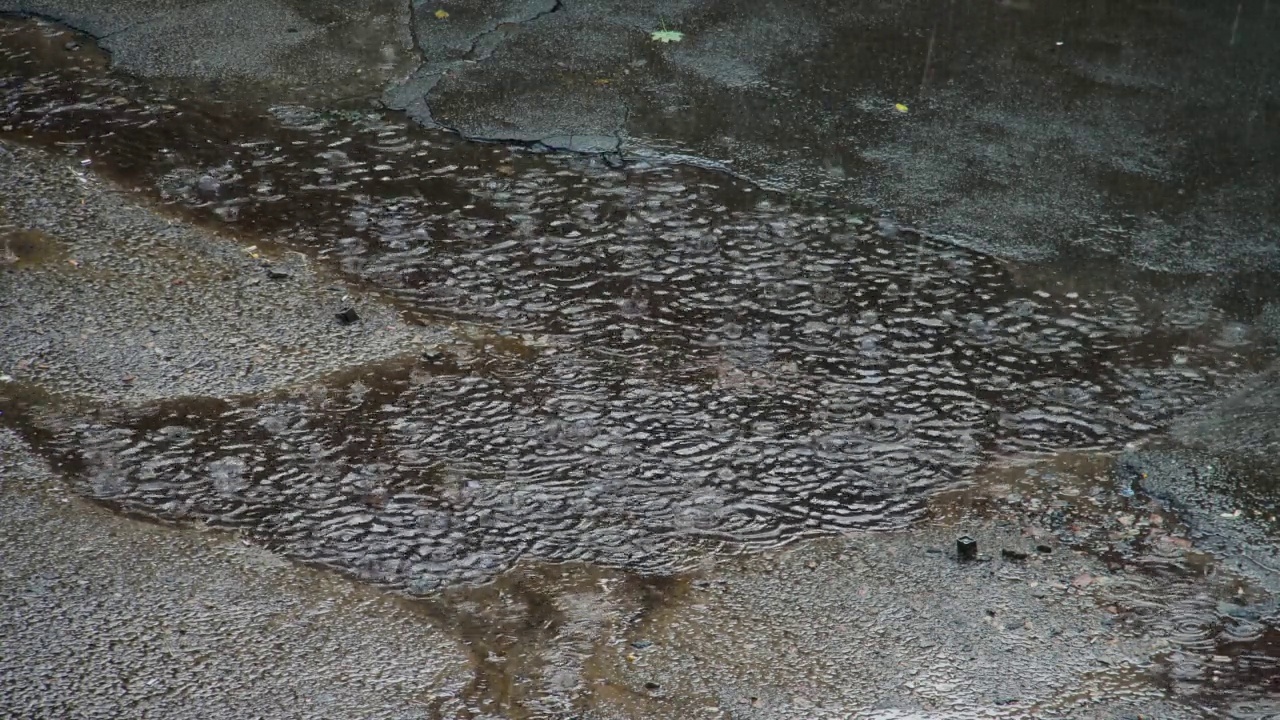 雨滴落入水坑。缓慢的运动。大雨滴落在柏油路上的水坑里，在水中散开成波。白天水中天空的倒影。大雨。恶劣的天气视频素材