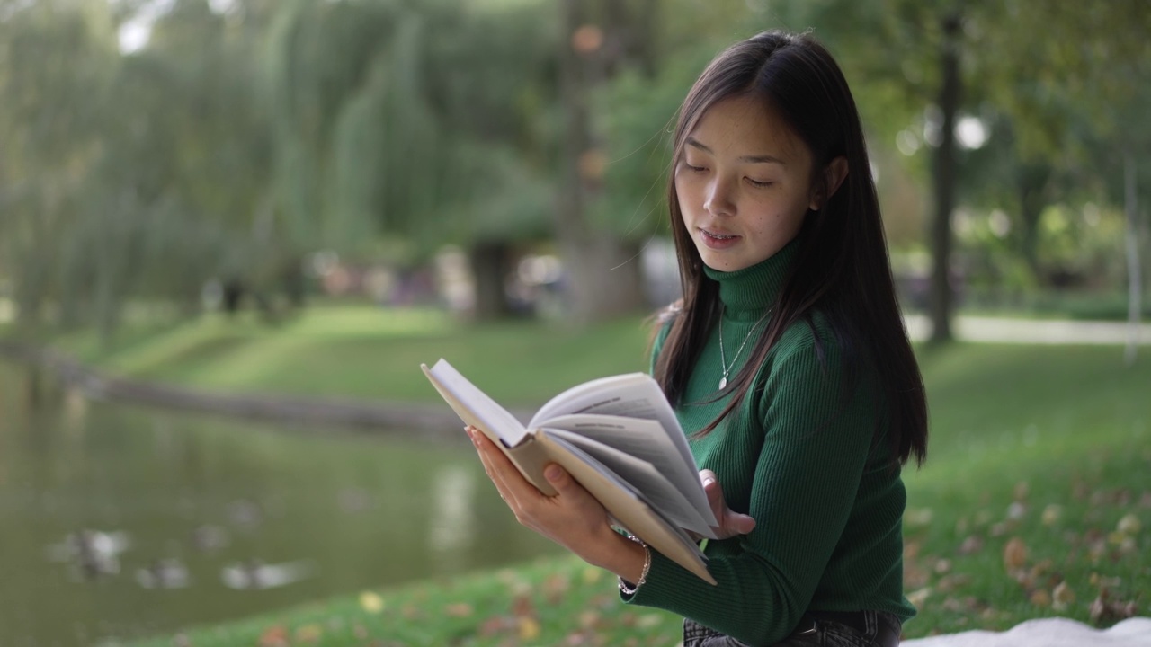 年轻的微笑聪明的女人在夏天的公园在池塘边翻页读书。肖像自信轻松快乐的亚洲女士享受户外的爱好在阳光灿烂的日子。生活方式的概念。视频素材