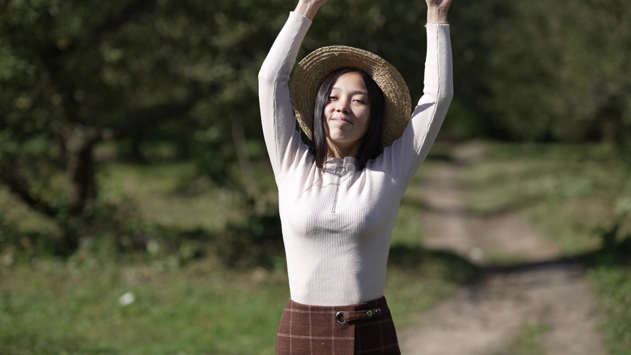 前视图微笑的妇女在草帽看着相机走慢动作在阳光明媚的公园离开。自信开朗的亚洲女士的肖像在阳光下散步。视频素材