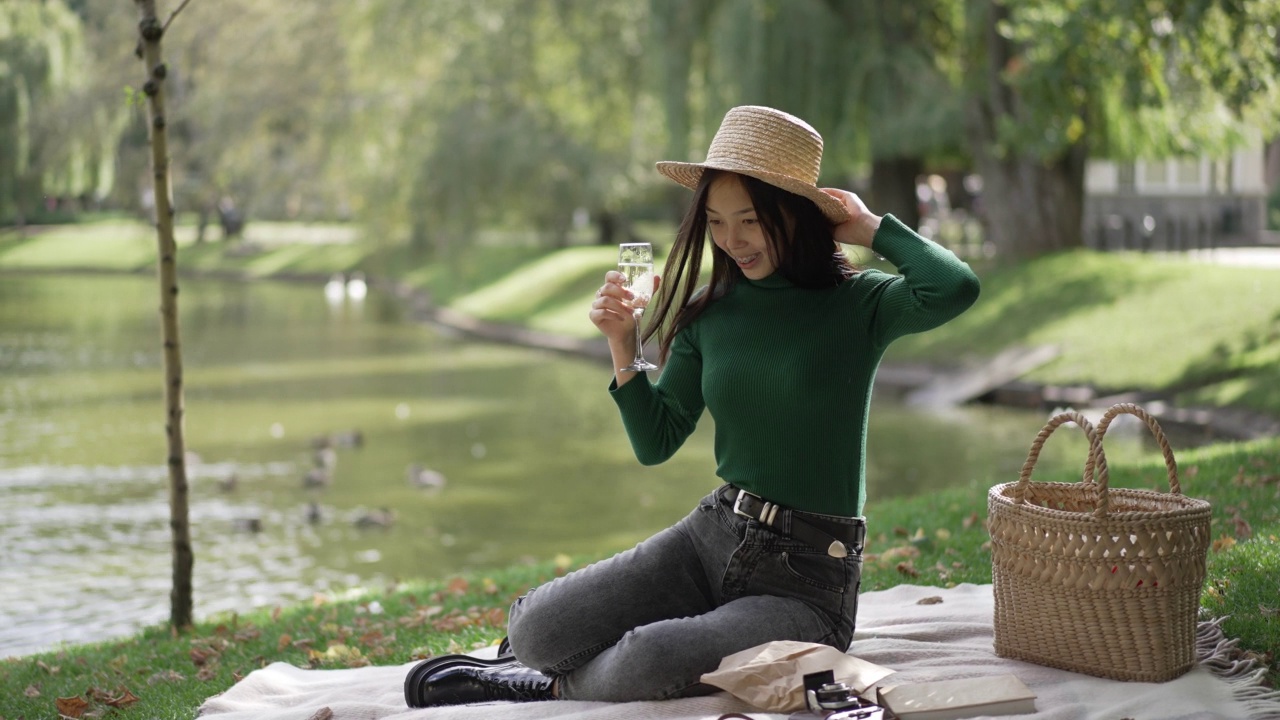 广角拍摄微笑的亚洲苗条年轻女子喝香槟杯野餐在夏季公园。肖像华丽自信迷人的女士在草帽享受饮料环顾四周。视频素材