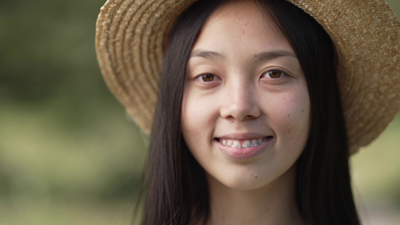 放大面对迷人的年轻女子牙套微笑看着相机。特写肖像亚洲苗条女士在草帽摆姿势慢动作户外。美丽和个性。视频素材