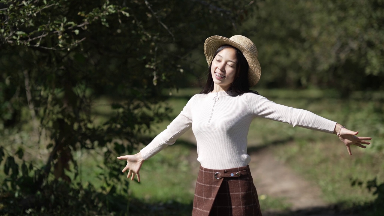 中拍肖像快乐的女人伸出手慢动作站在阳光室外。放松的亚洲千禧女士享受悠闲的阳光在夏季公园微笑看向别处。视频素材