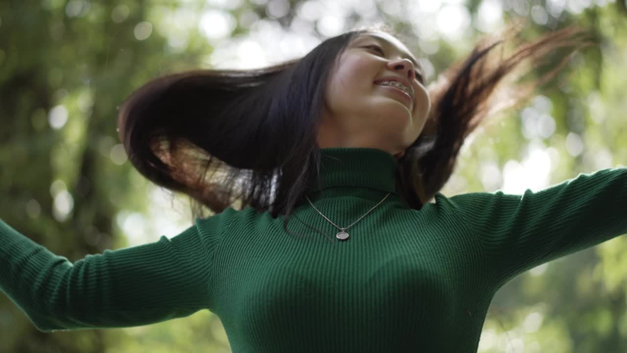 华丽的亚洲女人在阳光下旋转，微笑着看着镜头甩头发。画像的快乐无忧无虑的年轻苗条的女士摆姿势在慢动作享受休闲户外的阳光。视频素材