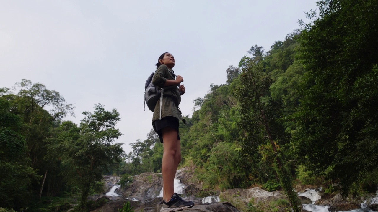 背着背包的旅行女子站在热带雨林的视点上，看着瀑布。视频素材