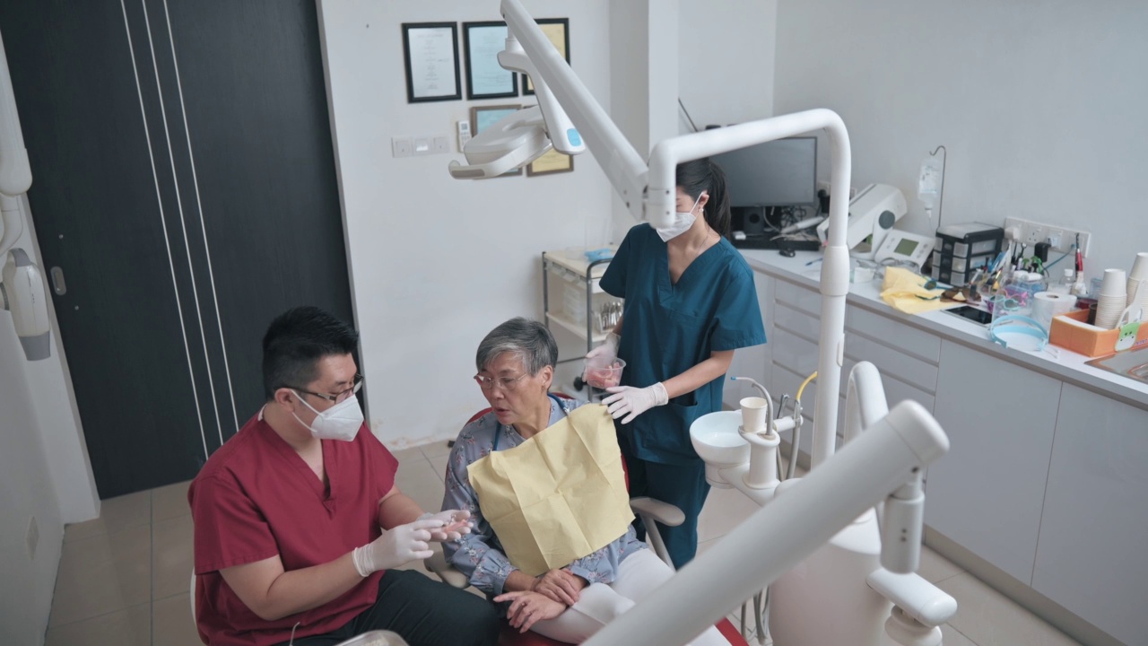 亚裔华人男性牙医展示假牙，向老年女性患者解释解决方案视频素材