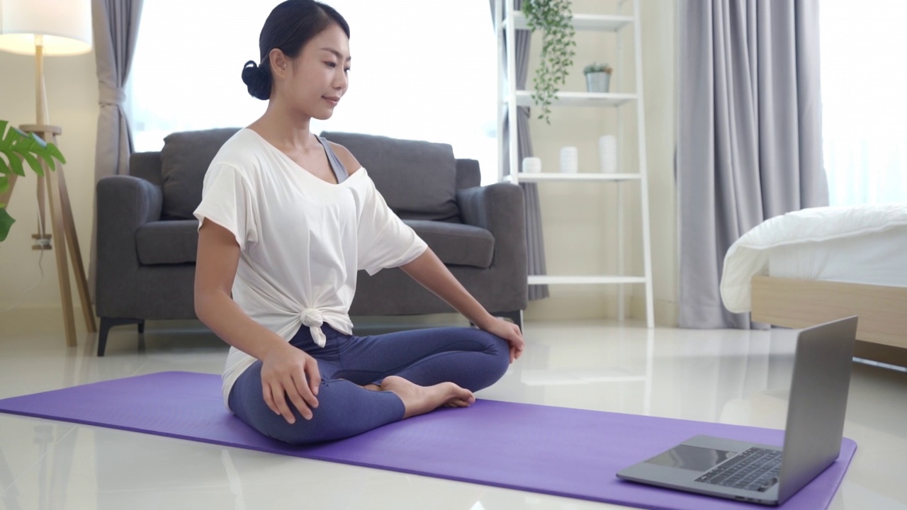 一位亚洲年轻女子正在开始锻炼。她试图通过笔记本电脑上的在线健身视频学习在瑜伽垫上做伸展运动。视频素材