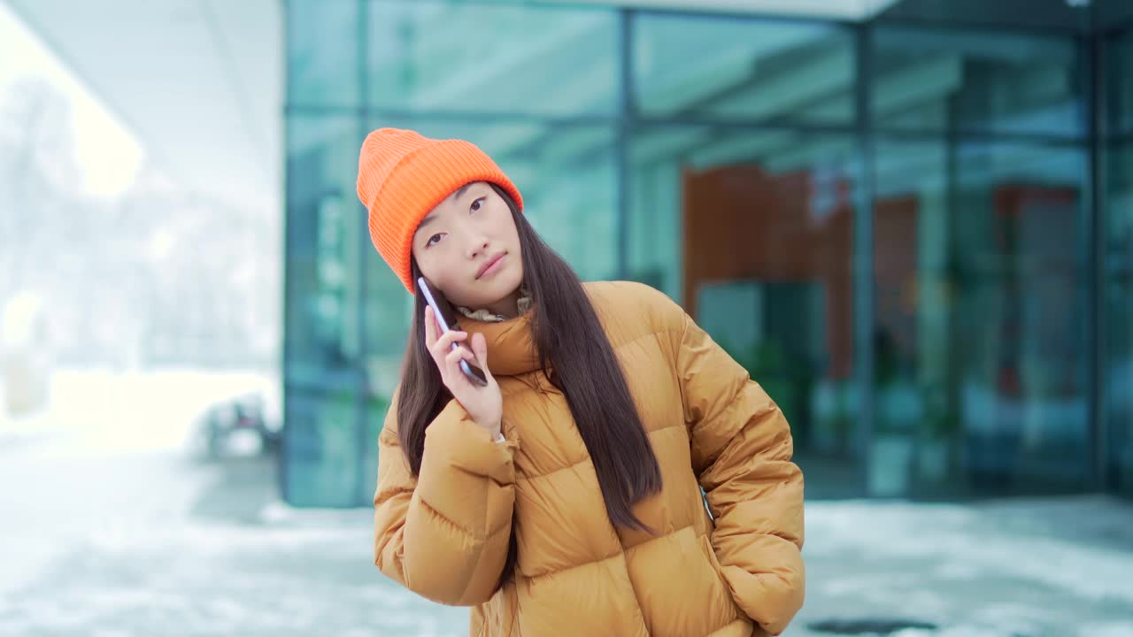 年轻的亚洲女性在一个下雪的冬日里打电话，她是在校园里的学生。在现代都市背景下，站在城市街道上快乐的女孩对话视频素材