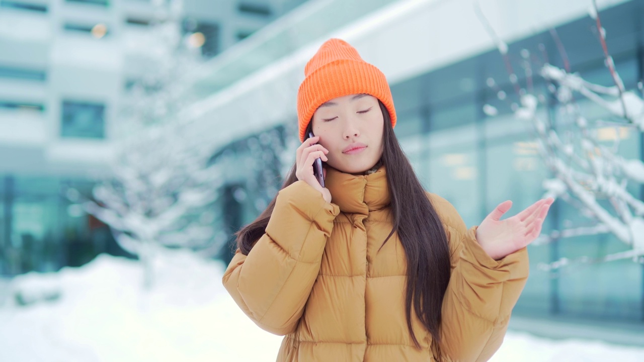 年轻的亚洲女性在一个下雪的冬日里打电话，她是在校园里的学生。在现代都市背景下，站在城市街道上快乐的女孩对话视频素材