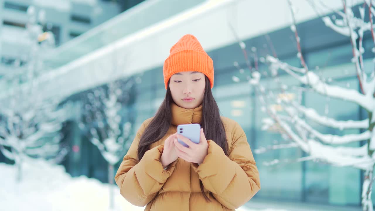 美丽快乐的亚洲少女学生站在冬雪城市街道在寒冷的天气使用手机微笑。年轻女子手里拿着智能手机滚动短信聊天使用。视频素材