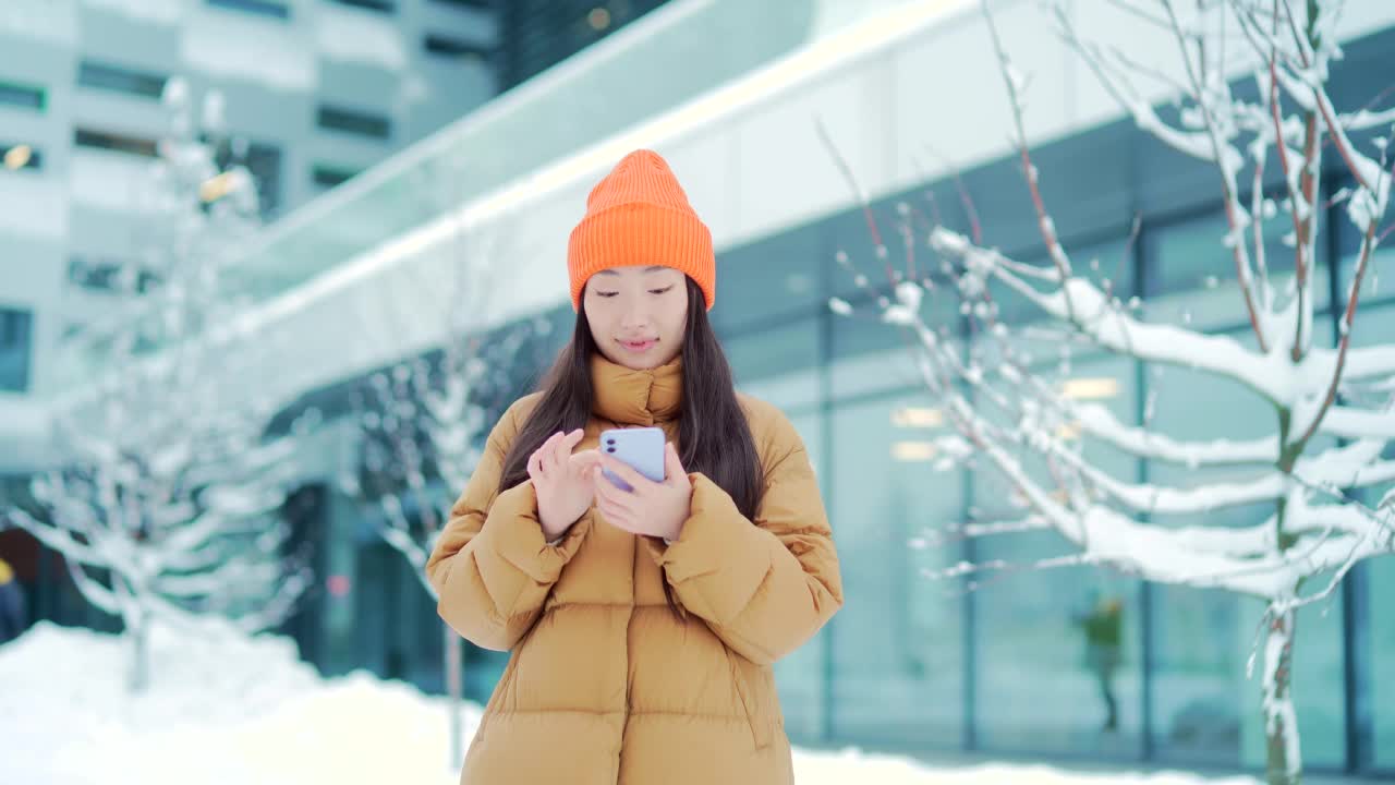 美丽快乐的亚洲少女学生站在冬雪城市街道在寒冷的天气使用手机微笑。年轻女子与智能手机在手中滚动短信聊天使用。视频素材