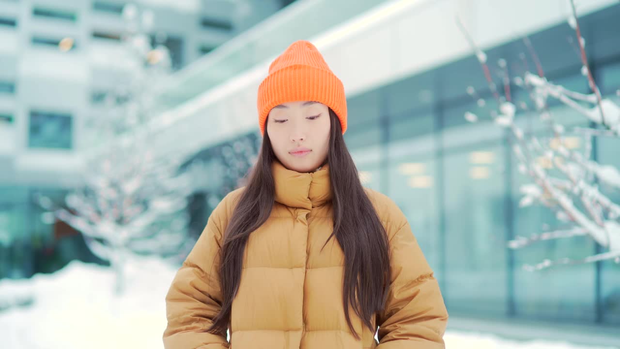 画像年轻的亚洲妇女站在雪地背景在现代城市街道建筑在寒冷的天气看着相机。面对快乐微笑的美丽女孩在冬天的户外视频素材